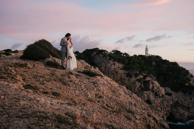 A couple standing on rust-coloured cliffs above the sea at sunset, a lighthouse behind them