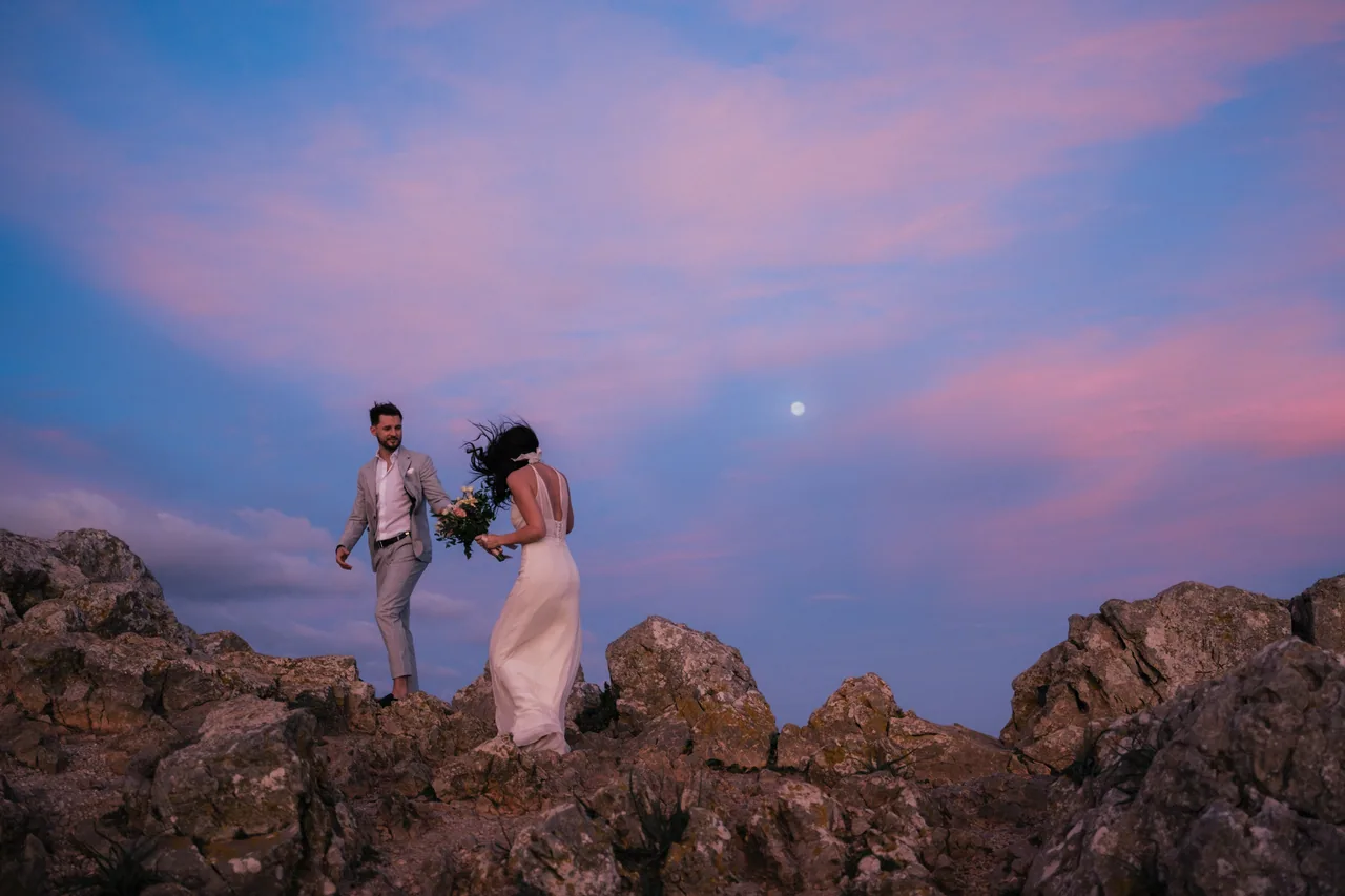 A couple standing on coastal rocks under a pink and violet sunset sky on Mallorca