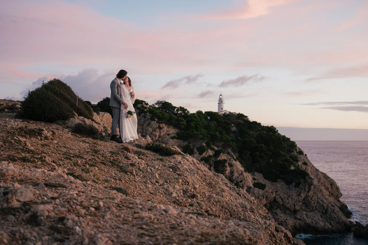 A couple on a clifftop above the sea with a distant lighthouse at dusk on Mallorca
