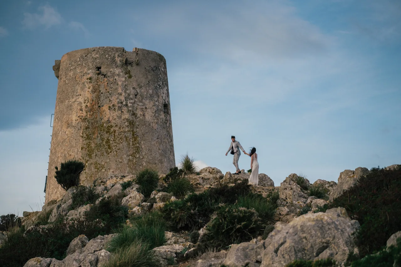 A couple walking on coastal rocks below an old stone watchtower on Mallorca